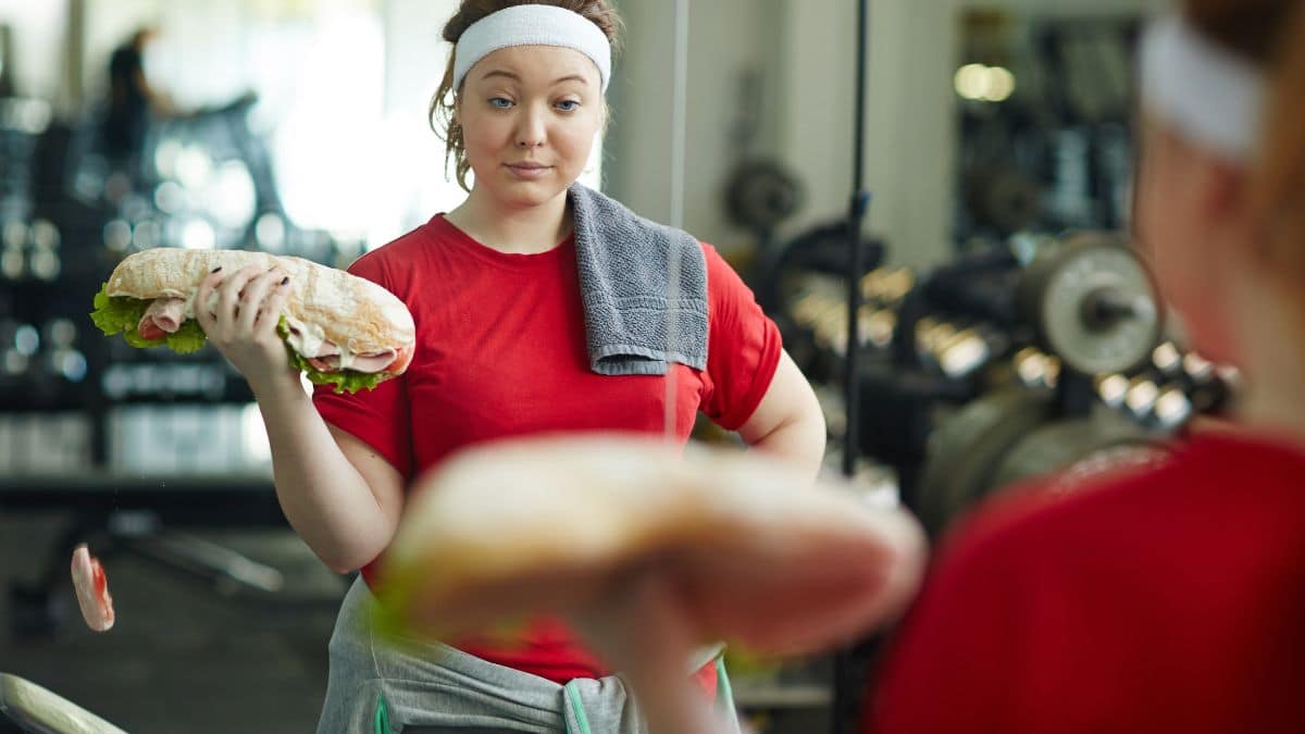 woman in gym wearing red shirt and white headband holding sandwich